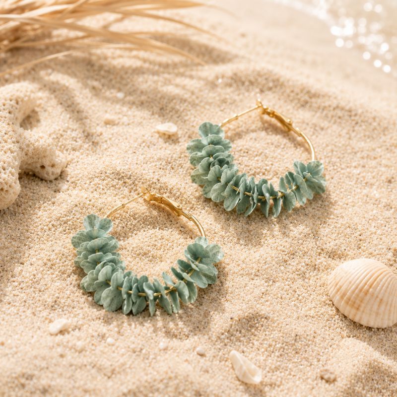 A pair of light blue leaf-shaped earrings on sand with shells and straw in the background