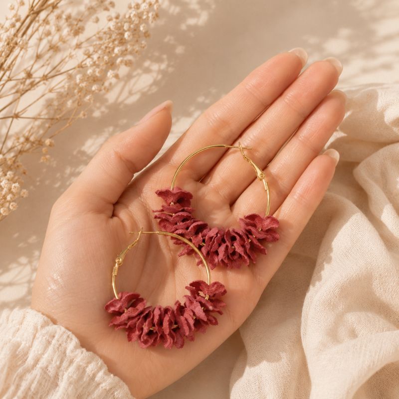 Hand holding gold hoop earrings with red floral charms against a soft beige background