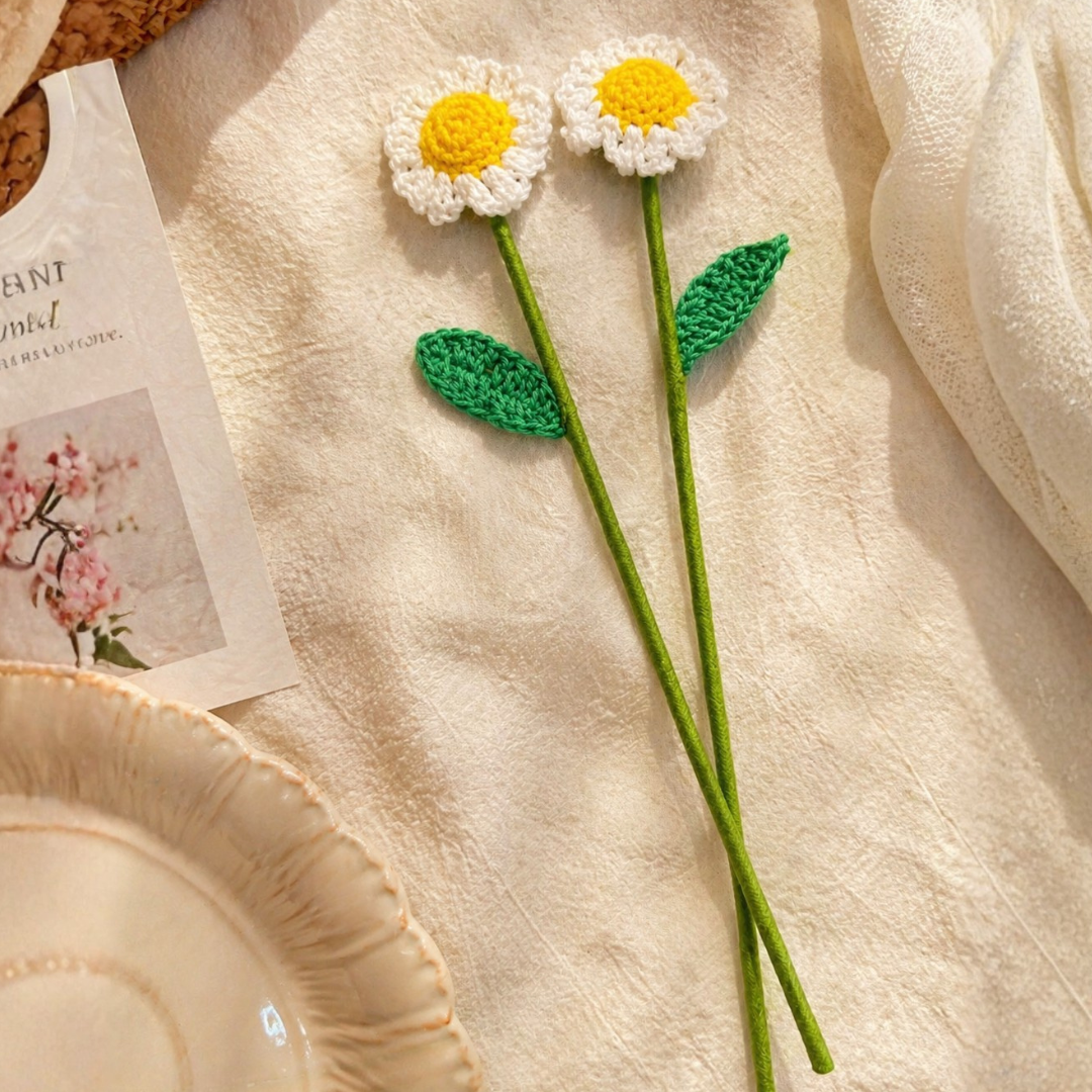 Two crocheted daisies with green stems and leaves on a textured beige surface.