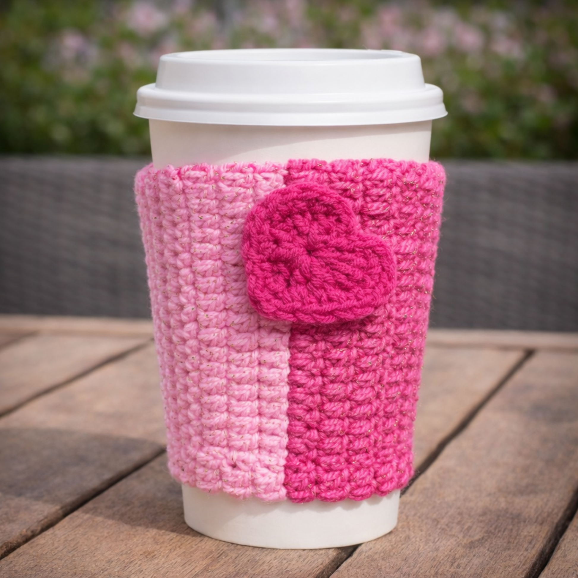 Coffee cup with a pink crochet sleeve featuring a flower on a wooden surface.