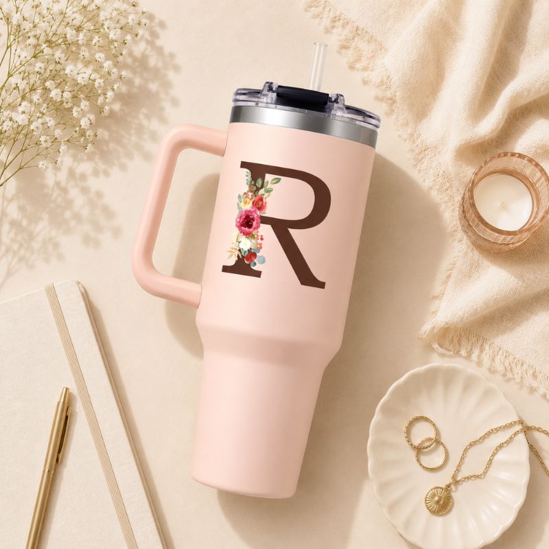 Pink tumbler with floral monogram on a beige surface with flowers and jewelry.
