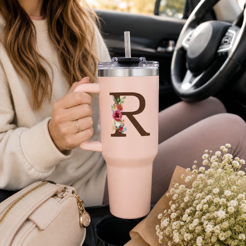 Person holding a pink tumbler with a floral 'R' design in a car.