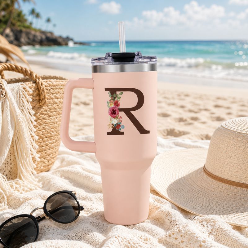 Pink tumbler with letter 'R' on a sandy beach with sunglasses and a hat.