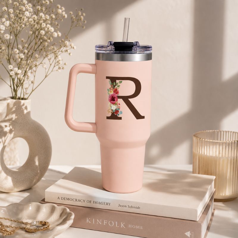 Pink tumbler with floral design and letter 'R' on a stack of books with decorative items around.