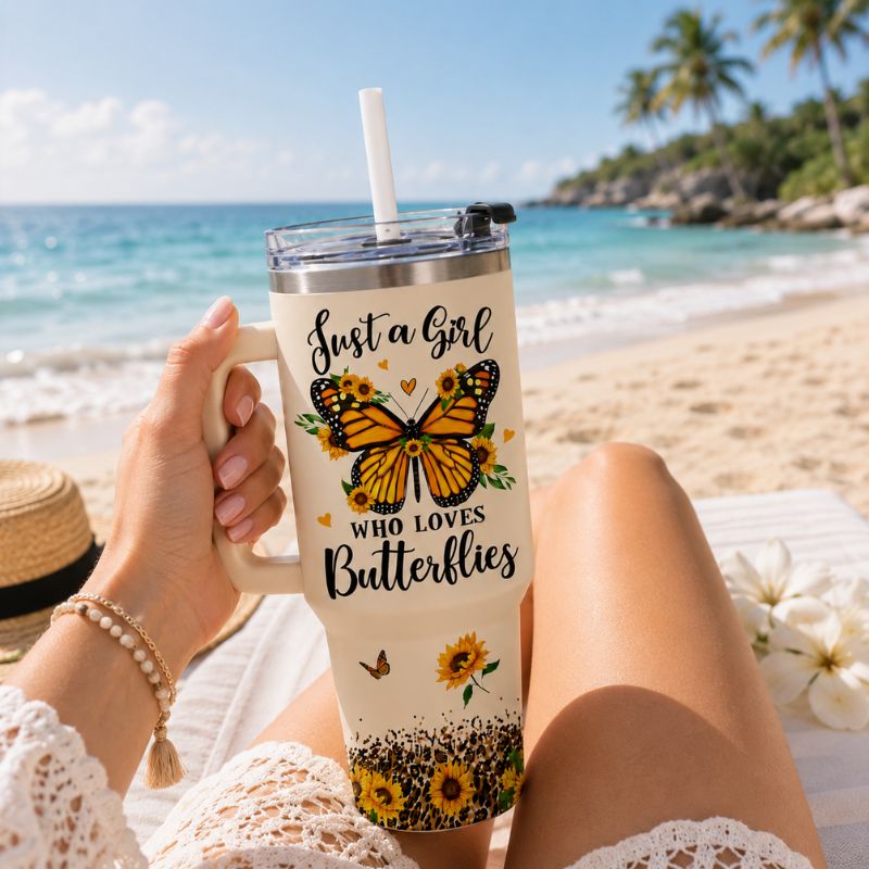 Person holding a tumbler with butterfly design and text on a beach
